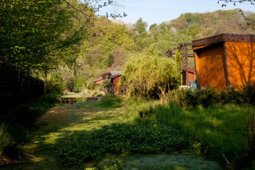 Ein üppiger grüner Garten mit einem Teich, verschiedenen Pflanzen und einer Reihe von hölzernen und orangefarbenen Gebäuden im Hintergrund, umgeben von Bäumen und Hügeln.