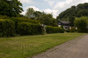 Ein Haus, das teilweise von hohen Büschen und Bäumen verdeckt ist, mit einem grasbewachsenen Rasen und einem gepflasterten Gehweg unter einem teilweise bewölkten Himmel.