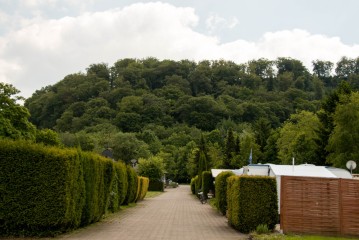 Ein gepflasterter Weg führt durch einen Campingplatz mit gestutzten Hecken, Zelten und Wohnwagen zu einem dicht bewaldeten Hügel unter einem teilweise bewölkten Himmel.