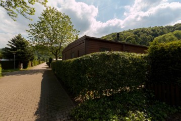 Ein Holzhaus mit Hecke und Zaun entlang eines gepflasterten Weges, umgeben von Bäumen und Grünpflanzen unter einem teilweise bewölkten Himmel.