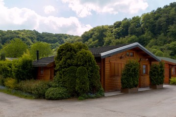 Eine Holzhütte mit geschlossenen Fensterläden ist von Sträuchern und Bäumen umgeben, vor einer Kulisse aus grünen Hügeln und einem teilweise bewölkten Himmel.