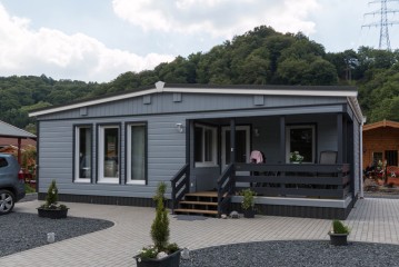 Ein modernes graues Haus in Modulbauweise mit einer überdachten Veranda, großen Fenstern und einer gepflasterten Auffahrt vor einer Kulisse aus grünen Bäumen und einem wolkenverhangenen Himmel.