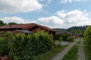 Eine Reihe von Holzhütten mit roten Dächern säumt einen Kies- und Grasweg, der unter dem teilweise bewölkten Himmel von grünen Hecken begrenzt wird.