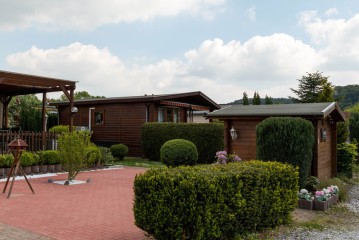 Ein kleiner, gepflegter Hof mit gestutzten Sträuchern, einem roten Ziegelweg und zwei Holzhäusern unter einem teilweise bewölkten Himmel.