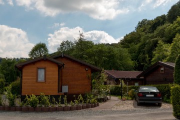 Einstöckige Holzhütten im Grünen, in der Nähe parkt ein schwarzes Auto unter einem teilweise bewölkten Himmel.