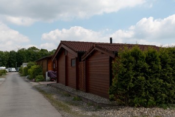 Eine Reihe brauner Holzhütten neben einer gepflasterten Straße, rechts Sträucher und Bäume, darüber ein bewölkter Himmel.