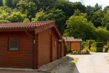An einem sonnigen Tag säumen Holzhütten mit roten Ziegeldächern eine gepflasterte Straße, umgeben von grünen Bäumen und Sträuchern.