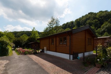 Eine kleine Holzhütte mit Schrägdach steht neben einer gepflasterten Auffahrt, umgeben von Grün und blühenden Sträuchern unter einem teilweise bewölkten Himmel.