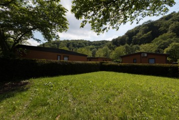 Drei einstöckige braune Gebäude sind von Hecken und Gras umgeben, mit Bäumen und bewaldeten Hügeln im Hintergrund unter einem teilweise bewölkten Himmel.