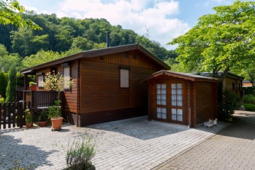 Ein kleines Holzhaus mit einem dazugehörigen Schuppen, umgeben von Bäumen und Topfpflanzen, steht auf einer gepflasterten Auffahrt unter einem teilweise bewölkten Himmel.