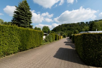 Ein gepflasterter Weg, gesäumt von hohen grünen Hecken, mit Bäumen und Hügeln im Hintergrund unter einem blauen Himmel mit Wolken.