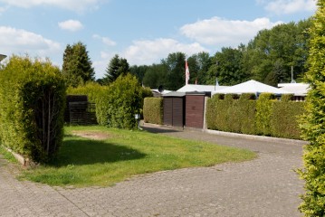 Gepflasterte Einfahrt mit Rasenfläche, umgeben von hohen grünen Hecken, die zu braunen Garagentoren und weißen Zelten im Hintergrund unter blauem Himmel führen.
