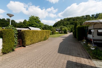 Ein gepflasterter Weg führt zwischen Hecken und geparkten Wohnwagen hindurch, mit grünen Bäumen und Hügeln im Hintergrund unter einem teilweise bewölkten Himmel.