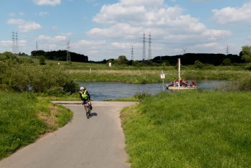 Ein Radfahrer fährt auf einem gepflasterten Weg in der Nähe eines Flusses, während im Hintergrund ein kleines Boot mit mehreren Passagieren auf dem Wasser zu sehen ist.
