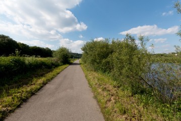 Gepflasterter Weg, umgeben von grünen Büschen und Gras, mit einem Fluss auf der rechten Seite unter einem blauen Himmel mit vereinzelten Wolken.