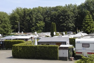 Reihen von Mobilheimen und Wohnwagen sind auf einem Campingplatz aufgestellt, der von gestutzten Hecken und hohen grünen Bäumen umgeben ist.