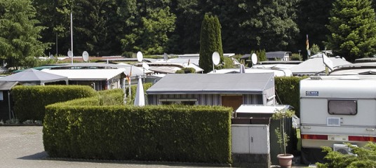 Reihen von Wohnwagen und kleinen Mobilheimen sind auf einem Campingplatz durch grüne Hecken voneinander getrennt, und auf vielen Dächern sind Satellitenschüsseln zu sehen.