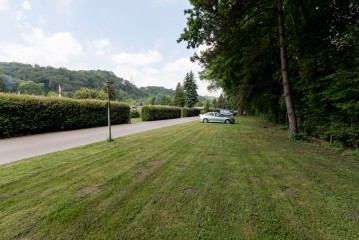 Ein helles Auto ist auf einer Wiese in der Nähe einer gepflasterten, von Hecken und Bäumen gesäumten Straße geparkt, mit einem bewaldeten Berghang im Hintergrund.