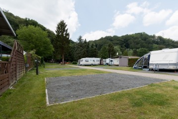 Ein Schotterparkplatz auf einem Campingplatz mit Wohnwagen, Grasflächen, Bäumen und Hügeln im Hintergrund unter einem teilweise bewölkten Himmel.