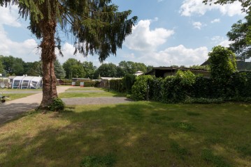 Eine Grasfläche mit einem hohen Baum im Vordergrund, einem gepflasterten Weg und Gebäuden, die von Grün umgeben sind, unter einem teilweise bewölkten Himmel.