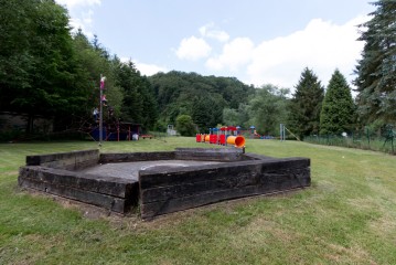 Ein Holzsandkasten ohne Sand in einem grasbewachsenen Park, mit Spielgeräten und Bäumen im Hintergrund.