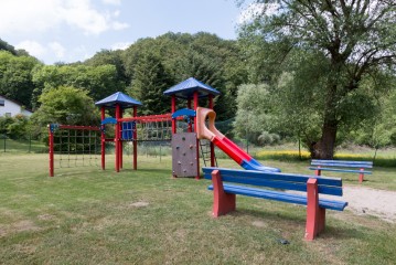 Ein Spielplatz mit Kletternetzen, einer Rutsche und blauen Bänken befindet sich auf einer Wiese mit Bäumen und Hügeln im Hintergrund.