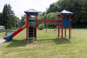 Ein Spielplatz mit einem roten und blauen Klettergerüst, einer Rutsche und einer Seilbrücke auf einer Wiese mit Bäumen im Hintergrund.