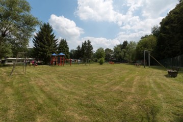 Eine grasbewachsene Wiese mit einem Spielplatz, Schaukeln, Rutschen und Bänken, umgeben von Bäumen unter einem teilweise bewölkten Himmel.