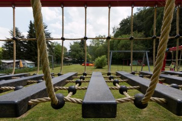 Nahaufnahme einer Seilbrücke und eines Klettergerüsts auf einem Spielplatz im Freien, mit Gras, Bäumen und Spielgeräten im Hintergrund.