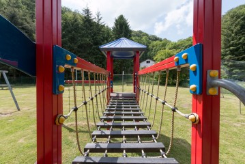 Eine Seilbrücke auf einem bunten Spielplatz Struktur, mit grünem Gras und Bäumen im Hintergrund unter einem teilweise bewölkten Himmel.