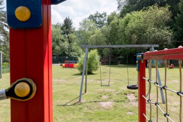 Ein grasbewachsener Spielplatz mit einer Schaukel, einem Kletternetz und einem rot-blauen Spielgerät, umgeben von Bäumen und Grünflächen.