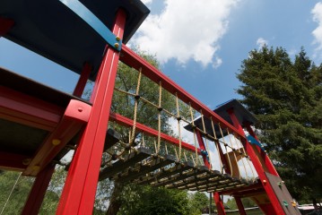 Klettergerüst auf dem Spielplatz mit roten Balken, Seilbrücke und Holzstufen, fotografiert aus einem niedrigen Winkel vor einem blauen Himmel mit Wolken und Bäumen im Hintergrund.
