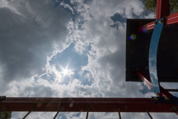 Blick auf einen wolkenverhangenen Himmel mit durchscheinender Sonne, gesehen von unten auf ein rot-blaues Spielplatzgebäude.