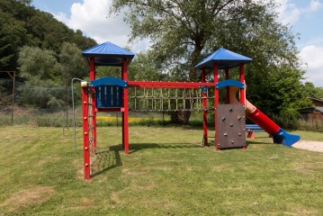 Eine bunte Spielplatzstruktur mit zwei Türmen, einer Seilbrücke, einer Kletterwand, einer Leiter und einer roten Rutsche steht auf einer Wiese mit Bäumen im Hintergrund.