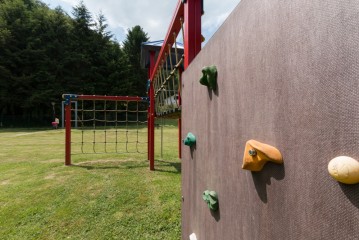 Im Vordergrund ist eine Spielplatz-Kletterwand mit farbigen Griffen zu sehen, im Hintergrund ein Seilnetz und grünes Gras.