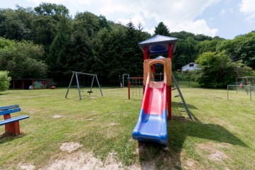 Ein Spielplatz mit Rutsche, Schaukeln, Klettergerüst und Bänken auf einer grasbewachsenen Wiese, umgeben von Bäumen und einem Haus im Hintergrund.