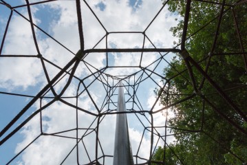 Blick nach oben durch eine netzartige Kletterstruktur aus Seilen mit einer Metallstange in der Mitte, vor einem teilweise bewölkten Himmel und belaubten Bäumen.