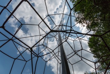 Blick nach oben durch eine Metallkletterstruktur mit geometrischen Formen, vor einem blauen Himmel mit Wolken und grünen Zweigen am Rande.