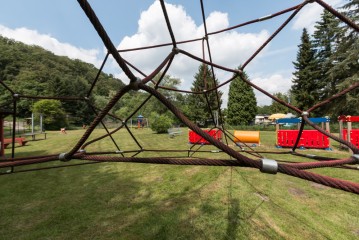 Ein Spielplatz mit Kletterseilen im Vordergrund, grünes Gras, Bäume, Bänke und verschiedene Spielgeräte im Hintergrund unter einem teilweise bewölkten Himmel.