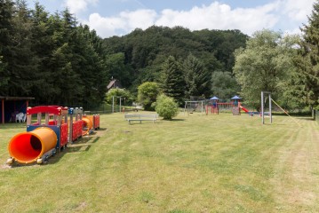 Ein grasbewachsener Spielplatz mit einem bunten Spielzug, Bänken, Schaukeln und Rutschen, umgeben von Bäumen und einem bewaldeten Hügel im Hintergrund.