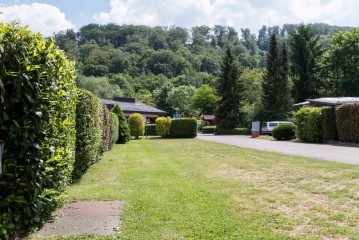 Eine gepflegte, von Hecken und Bäumen gesäumte Rasenfläche mit einer gepflasterten Straße und mehreren Häusern oder Hütten an einem bewaldeten Hang unter einem teilweise bewölkten Himmel.