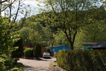 Ein Auto parkt auf einer gepflasterten Auffahrt zwischen grünen Hecken, Bäumen und mehreren kleinen Häusern, mit einem bewaldeten Hügel im Hintergrund.