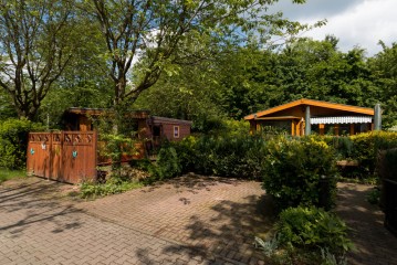Eine gepflasterte Zufahrt führt zu zwei kleinen Holzhütten, die von Bäumen, Sträuchern und Gartenpflanzen umgeben sind und unter einem teilweise bewölkten Himmel liegen.