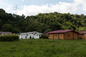 Ein grasbewachsenes Feld mit einem weißen Haus und einer Holzhütte vor einem bewaldeten Hügel. Im Hintergrund sind Stromleitungen und -türme bei teilweise bewölktem Himmel zu sehen.