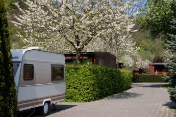 Ein weißer Wohnwagen steht neben einer Hecke an einer asphaltierten Straße, im Hintergrund blühende Bäume und Holzhütten.
