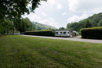 Ein weißer Wohnwagen ist in der Nähe einer Hecke neben einer asphaltierten Straße geparkt; Grasflächen und Bäume umgeben die Szene unter einem teilweise bewölkten Himmel.