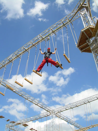 Eine Person, die eine Sicherheitsausrüstung trägt, navigiert durch einen Hochseilgarten mit Plattformen und Seilen, der an Gurten aufgehängt ist, vor einem blauen Himmel mit vereinzelten Wolken.