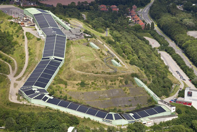Luftaufnahme einer großen Solaranlage auf den Dächern von Gebäuden, die sich an einem grasbewachsenen Hang neben einer Straße und bewaldeten Flächen befinden.