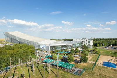 Eine große überdachte Skipistenanlage mit Glasfassade, umgeben von Bäumen, Erlebnisparcours und Sitzgelegenheiten im Freien unter blauem Himmel.