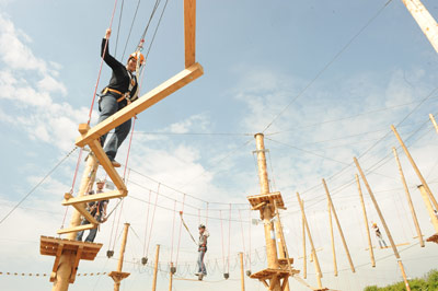 In einem Hochseilgarten unter blauem Himmel bewegen sich die Teilnehmer mit Klettergurten über Holzplattformen und Seilbrücken.